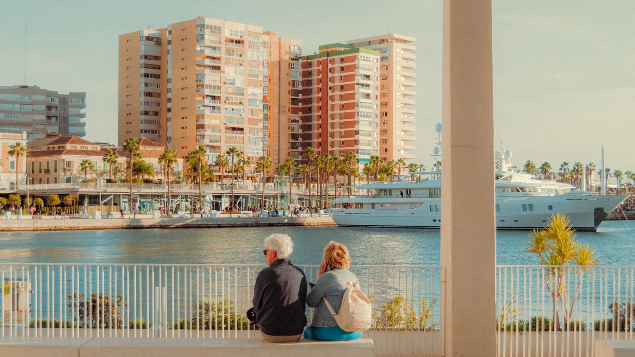 A senior couple enjoys the view by the marina in Málaga, Spain, with urban buildings in the backdrop.